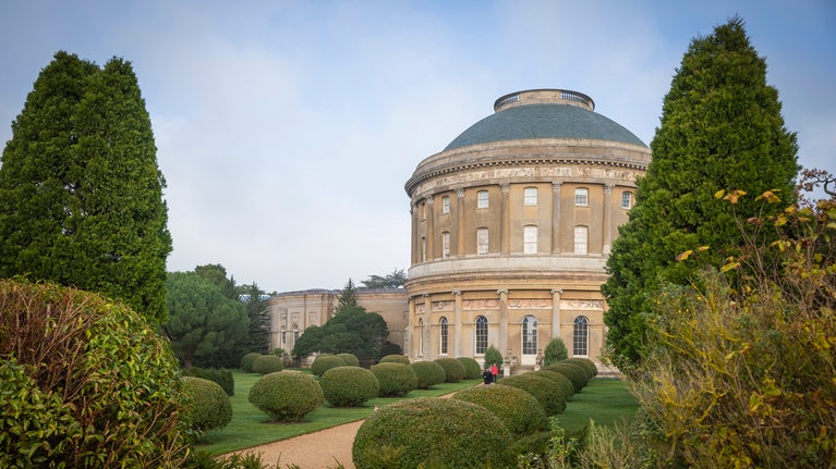 The rotunda is right of centre and framed by a tree on the far left and right of the image. in front is a path leading towards the left lined by round trimmed bushes